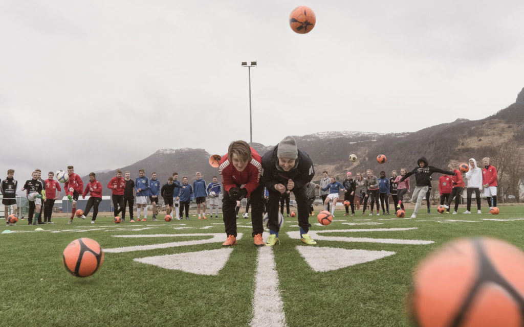 To ledere bøyer seg ned på en fotballbane mens 30 ungdommer forsøker å sparke fotballer på dem