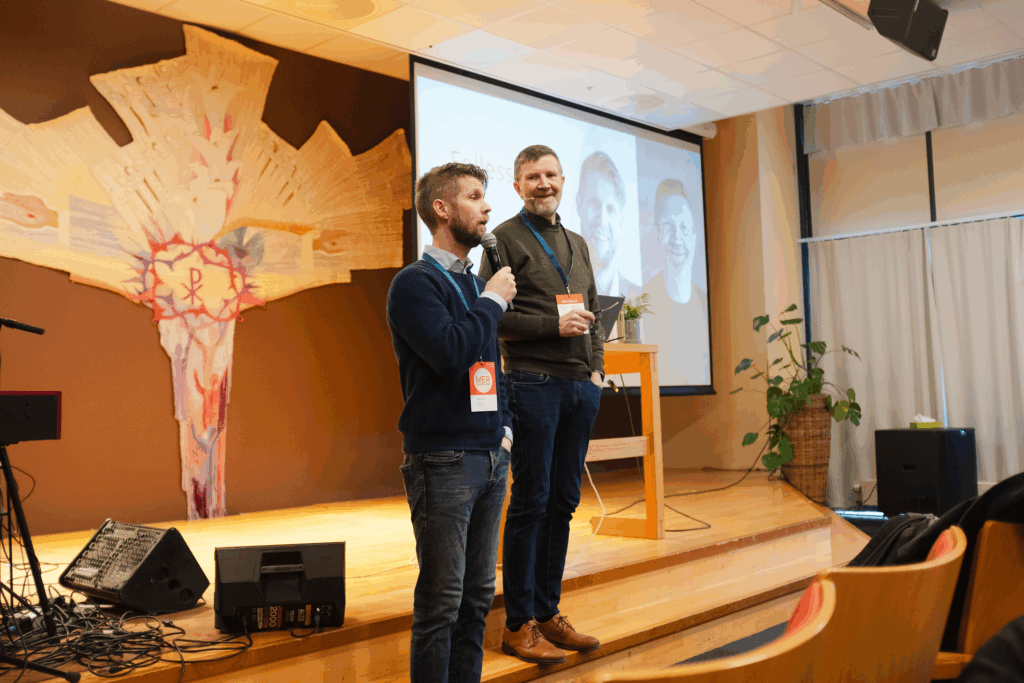 To menn står på en scene. Den ene snakker i mikrofonen